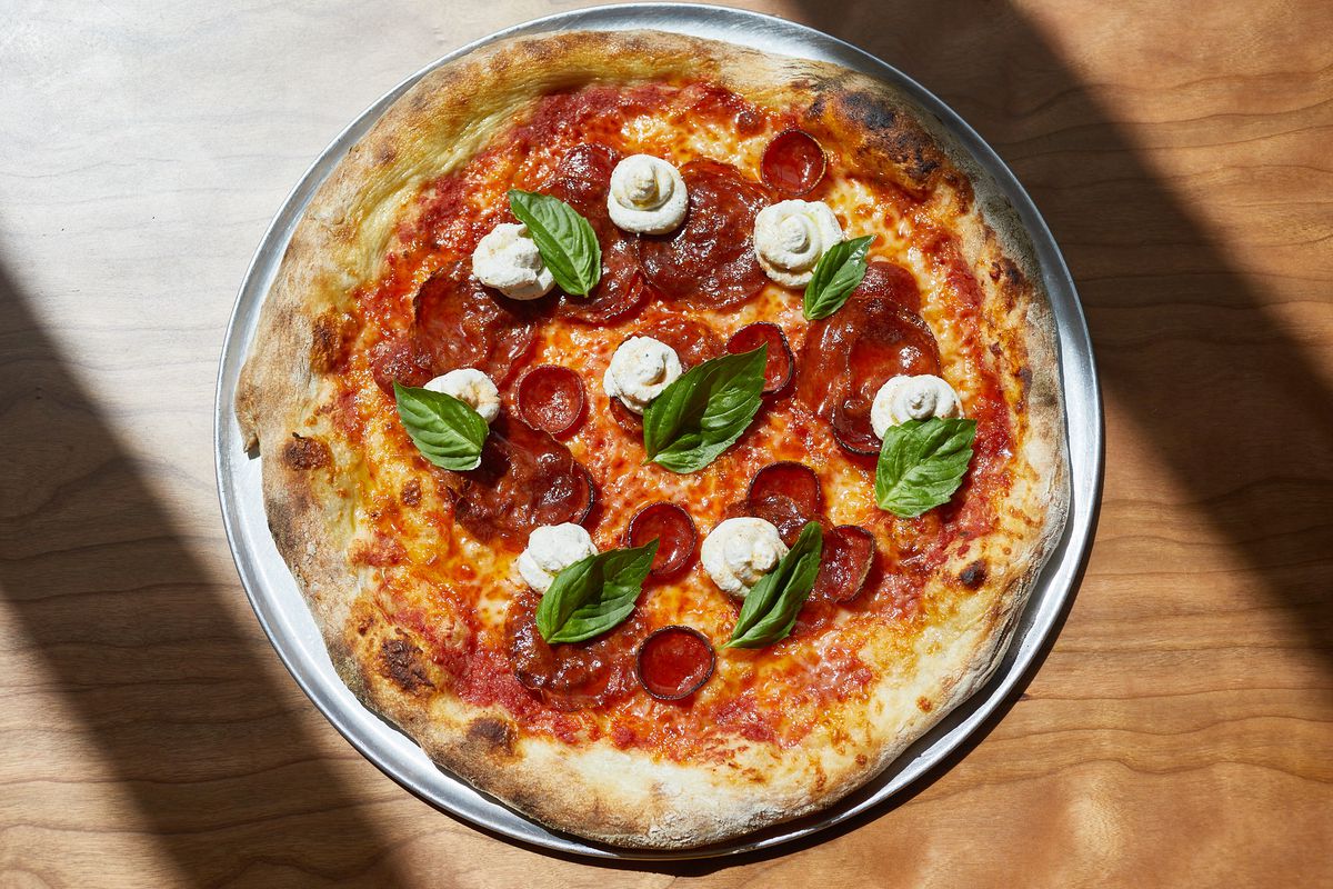 Pizza bundar dengan pepperoni, daun kemangi, keju putih, di atas meja.