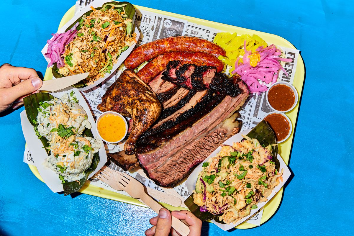 A yellow tray topped with barbecue, sauces, and sides — including ribs, jalepeño pork sausage, and smoked chicken, as well as pickled onions, sliced banana peppers, rujak slaw, potato salad, and nasi goreng.