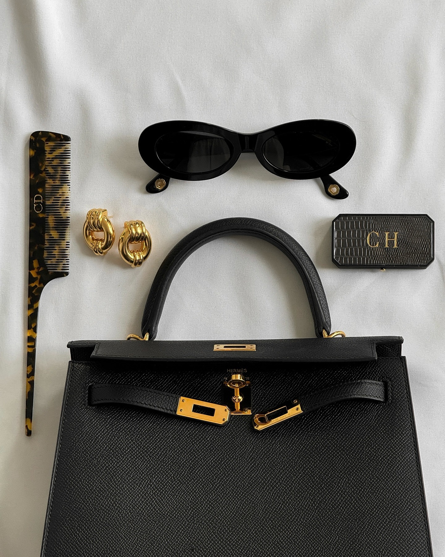 Foto tas tangan Hermes Kelly hitam, sisir kulit penyu, anting emas, kacamata hitam. dan kotak lip balm bermotif inisial CH.